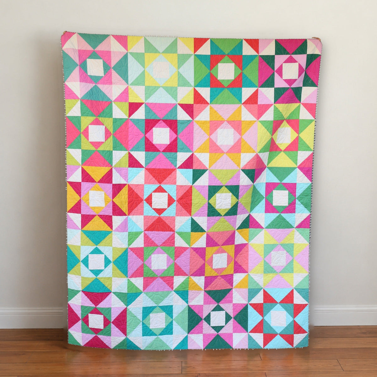 Bright scrappy Economy Stars quilt leaning against a cream wall on a wood floor