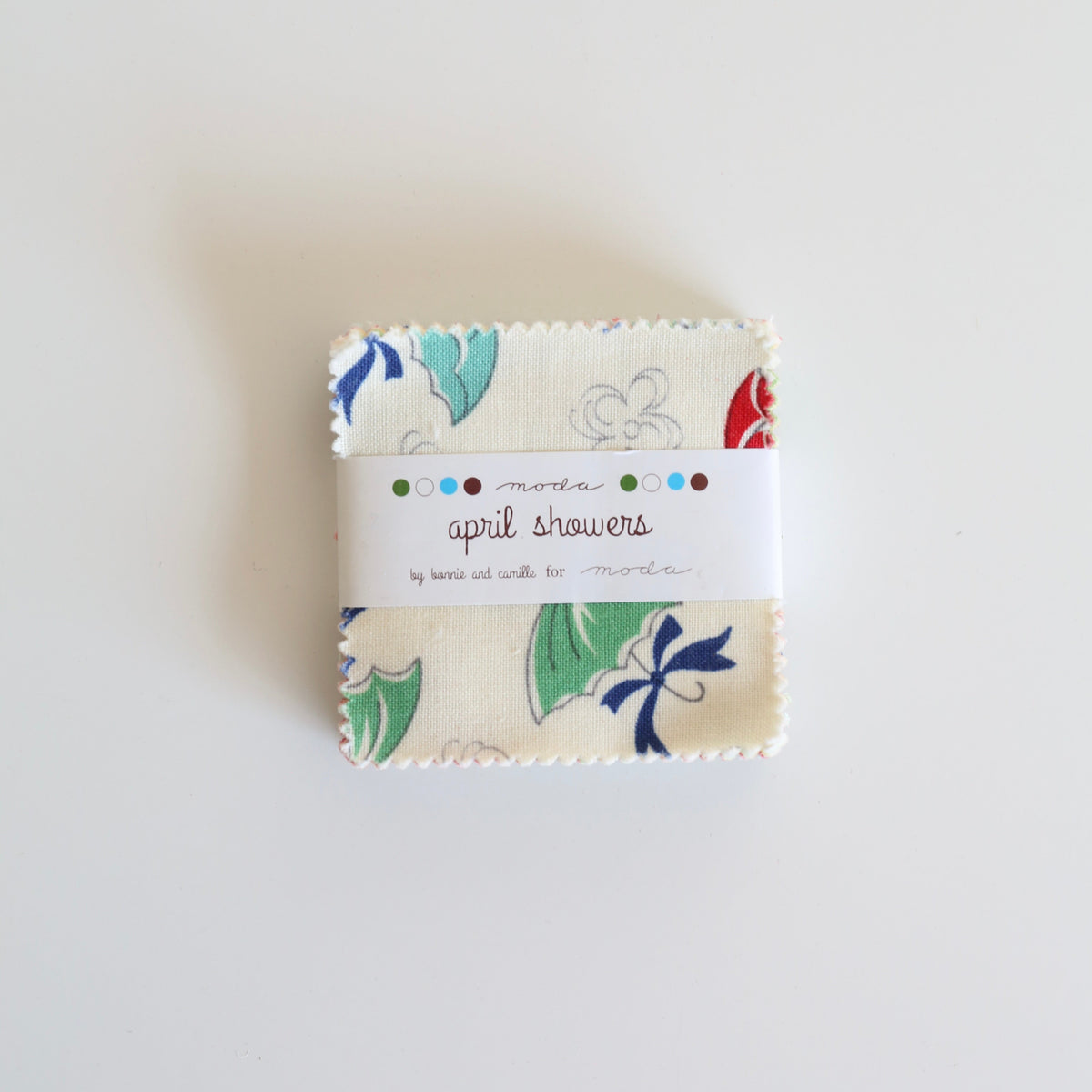 A small fabric bundle with a white paper label reading 'April showers' and 'by Jennifer Annitta' is centered on a plain white background. The fabric has a cream base with a multicolor floral and bird print in blue, green, red, and white.