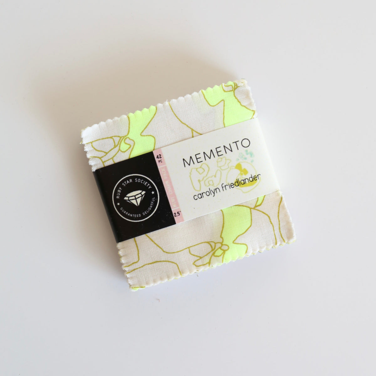 A small folded fabric bundle wrapped in a white paper band labeled 'MEMENTO' and 'carolyn freeman designer', with a black circular logo patch, photographed on a plain white background.