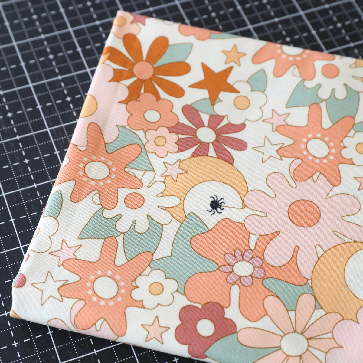A folded piece of fabric with a retro 70s floral pattern in orange, pink, and mint green on a cream background, placed on a black cutting mat.