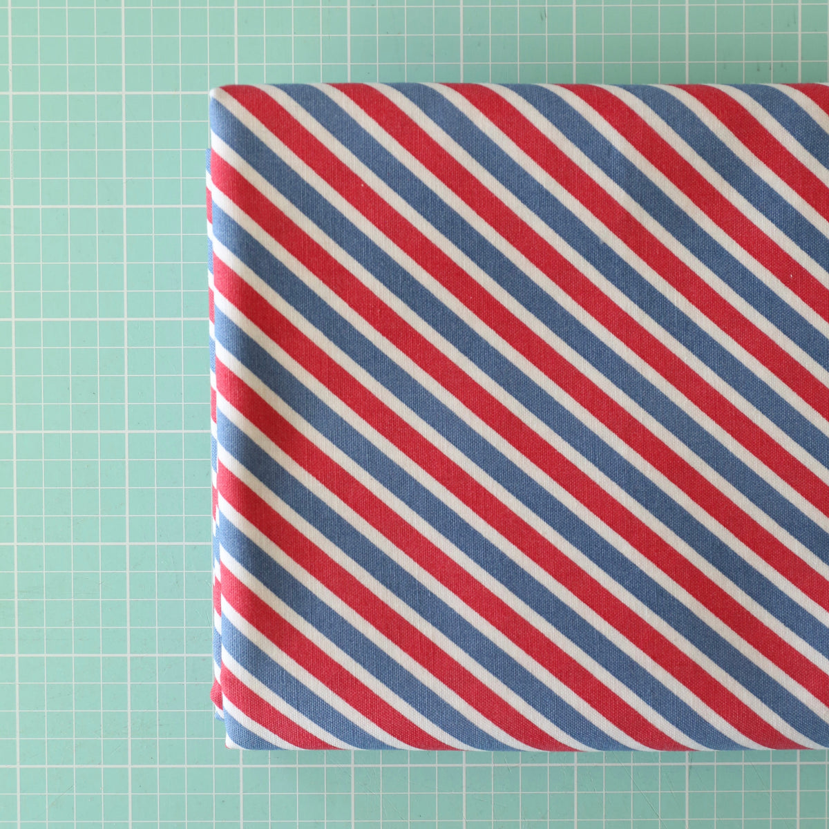 A folded piece of fabric with diagonal red, white, and blue stripes, placed on a mint green cutting mat with grid lines.