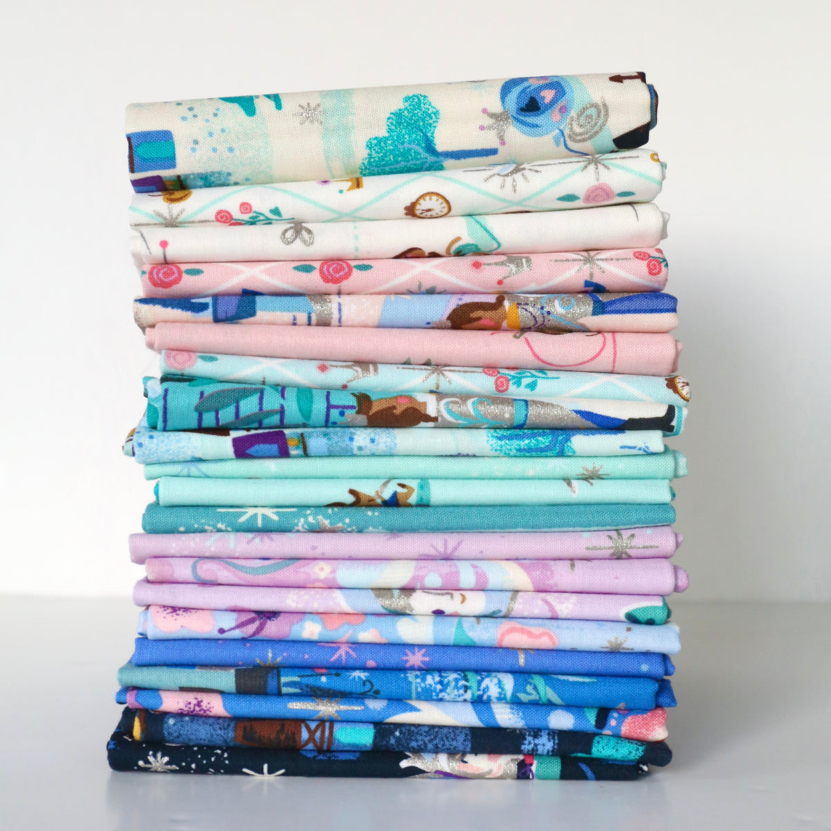 A tall stack of folded patterned fabrics in pastel and bright colors, including blues, pinks, purples, and whites, photographed against a plain light gray background.
